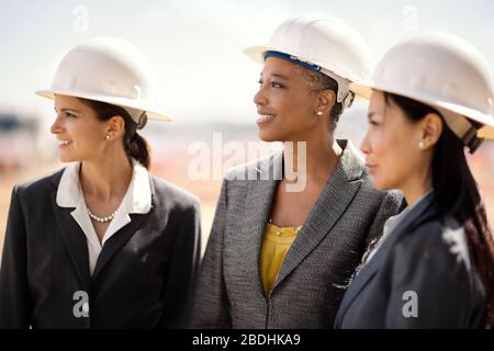 Drei lächelnde weibliche Kollegen, die Business-Anzüge und Hardhats tragen Stockfoto