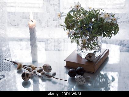 Eine Vase mit einem Blumenstrauß aus Frühlingsblumen steht auf einem alten Buch, neben Wachteleiern, Blumen und Weidenästen. Stockfoto