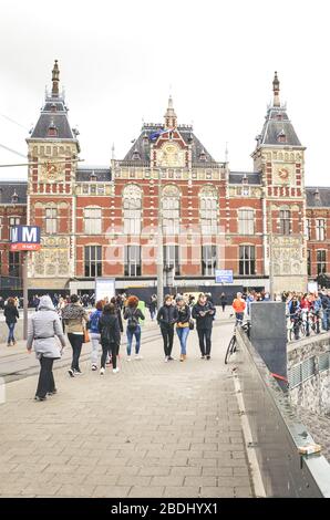 Amsterdam, Niederlande - 27. April 2019: Menschen in den Straßen der niederländischen Hauptstadt. Amsterdam Centraal, die Gebäude des Hauptbahnhofs im Hintergrund. Stadtzentrum, Kanal. Übergiebeltem Himmel. Stockfoto