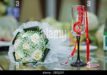 Die Ringe hingen von einem roten Band. Im Hintergrund ein Blumenstrauß für die Hochzeit. Im Glas sind Ringe und Bänder angebracht. Zwei Hochzeitsringe hingen von A. Stockfoto