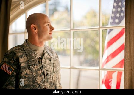 Nachdenklicher Soldat, der durch ein Fenster schaut, während er in seinem Haus steht. Stockfoto