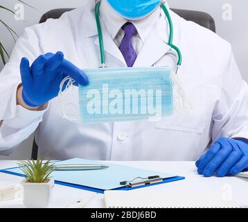 Der Arzt in einem weißen Mantel und blaue Latexhandschuhe sitzt an einem weißen Schreibtisch und zeigt einen Stapel medizinischer Einwegmasken, Schutzmittel gegen Viren und Stockfoto