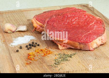 Rohes Fleisch auf einem Holztisch mit Knoblauch, Salz, schwarzem Pfeffer, Safran und Rosmarin zum Kochen bereit. Stockfoto