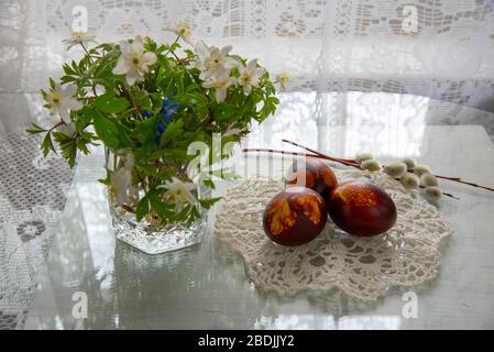 Ein Blumenstrauß aus Frühlingsblumen in einem Glas, bemalte Ostereier an einer gestrickten Serviette vor dem Hintergrund weißer Spitzen-Vorhänge. Stockfoto
