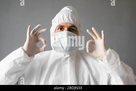 Arzt in weißem Schutzanzug, medizinischer Maske und Gummihandschuhen zeigt, dass alles in Ordnung ist. Hilfe für Menschen während der Bedrohung durch Coronavirus durch Pandemie Stockfoto