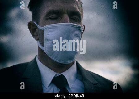Dunkles Porträt des Geschäftsmannes trägt chirurgische Gesichtsmaske im Freien mit geschlossenen Augen unter grauen Wolken Stockfoto