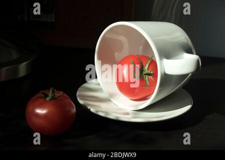 Zwei rote Tomaten in einer Tasse und auf einer weißen Platte in einem Sonnenstrahl auf einer schwarzen Tischoberfläche. Stockfoto
