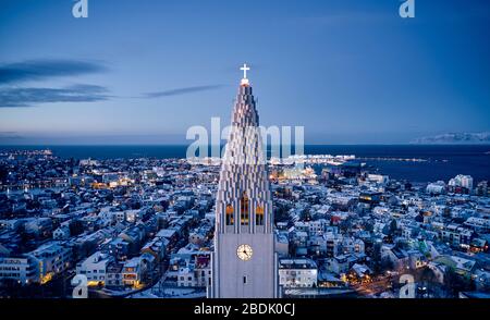 Drohnenblick auf den weißen hohen Turm der Hallgrimskirkja-Kirche mit glühendem Kreuz gegen die beleuchtete Stadt Reykjavik in der Abenddämmerung Stockfoto