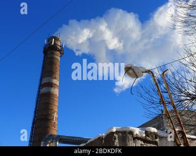 Rohre einer alten Fabrik werfen Wolken von giftigem weißem Rauch in den Himmel, die die Atmosphäre schädigen. Urbaner Smog aus Rauch aus Kesselhäusern. Weißer Rauch aus einem Kamin gegen einen blauen klaren Himmel. Stockfoto