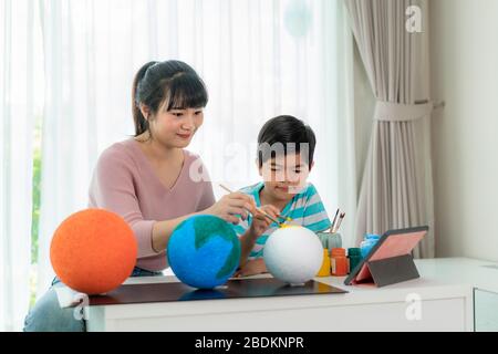 Asiatische Grundschüler mit Mutter malen den Mond in der Wissenschaftsklasse und lernen über das Sonnensystem per Videokonferenz mit Lehrer und anderem Stockfoto