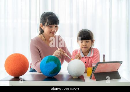 Asiatische Grundschulmädchen mit Mutter malen den Mond in der Wissenschaftsklasse und lernen über das Sonnensystem per Videokonferenz mit Lehrerin und anderem Stockfoto