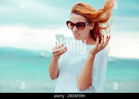 Porträt der gestressten jungen Frau, die das Handy in den Händen hält, die mit einem Gesichtsausdruck auf den Bildschirm blickt, verrückt nach stressigen Texten und Anrufen, die isoliert waren Stockfoto