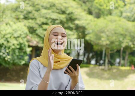 Porträt eines fröhlichen muslimischen Mädchens mit Smartphone, lächelnd im Freien. Stockfoto