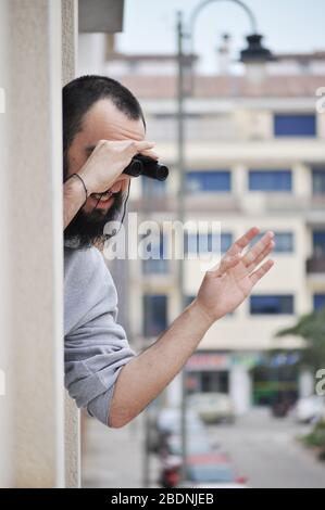 Der junge Mann winkt der Familie, während er sich aus dem Fenster der Eigentumswohnung auslehnt, das durch das Fernglas blickt. Mann beobachtet mit Fernglas in Spani durch Fenster Stockfoto