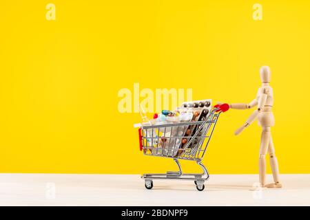 Holzpuppe rollt Metall Warenkorb mit Pillen auf gelbem Hintergrund. Pillen, Tabletten und Medikamente auf gelbem Hintergrund. Gesundheitskonzept. Stockfoto