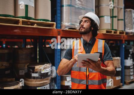 Schöner junger bärtiger Mann trägt Sicherheitsweste und Schutzhelm mit digitalen Tablet Eingabe von Daten in Lager stehen gegen Regal mit Karton Stockfoto