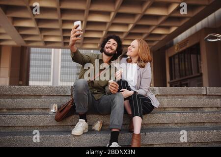 Fröhliches junges Paar in formeller Kleidung, das Selfie mit dem Smartphone nimmt, während es in der Pause zum Mittagessen ein Sandwich isst Stockfoto