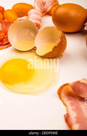 Zerbrochenes Hühnerei, Speck- und Wurstscheiben sowie Tomaten auf weißem Hintergrund. Nahaufnahme Stockfoto