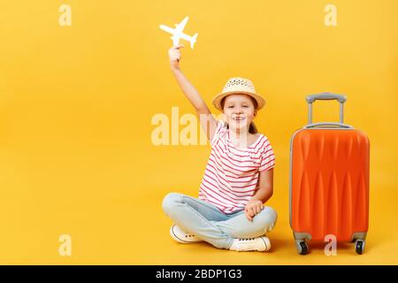 Fröhliches positiv schönes charmantes kleines Mädchen, das im Studio sitzt und ein Papierflugzeug hält und von Reisen träumt. Kind mit Koffer Stockfoto