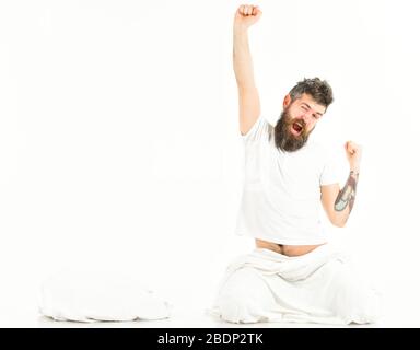 Mann streckt und gähnelt, wacht auf, sitzt auf dem Boden. Tätowierte Männer mit Bart Strecken sich die Arme, während sie morgens in der Nähe eines Kopfkissens sitzen, oben, Blick Stockfoto