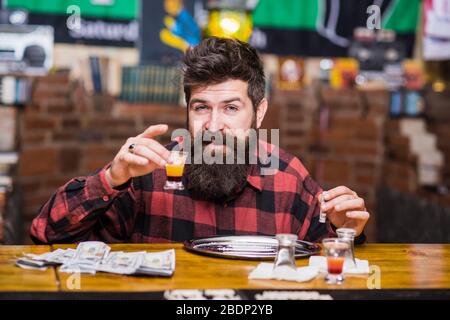 Hipster hält Shot-Cocktail und Röhrchen zum Schnüffeln von Drogen. Guy verbringen Freizeit in der Bar, die einen hohen, entschärften Hintergrund erhält. Illegales Spaßkonzept. Mann mit betrunkenem fröhlichem Gesicht allein an der Theke sitzen. Stockfoto