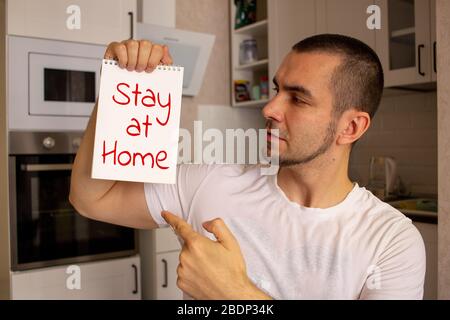 Während der COVID-19-Sperre bleibt ein junger Mann ohne Maske zu Hause. Halten Papier zu Hause bleiben Text. Stockfoto