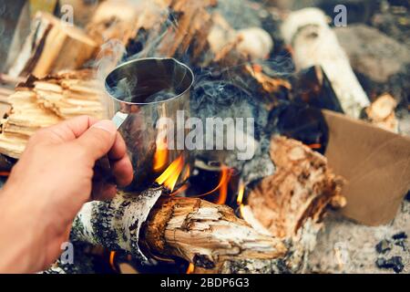 Man Traveler Hände halten Tasse Tee in der Nähe des Feuers draußen. Wanderer trinkt im Camp Tee aus dem Becher. Kaffee gekocht über einem Lagerfeuer auf der Natur Stockfoto