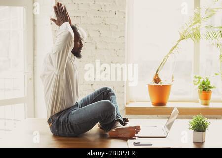 Junger afroamerikanischer Mann, der zu Hause Yoga macht, während er unter Quarantäne steht und freiberuflich online arbeitet. Remote, isoliert oder allein im Büro. Konzept des gesunden Lebensstils, Wellness, Aktivität, Bewegung. Stockfoto