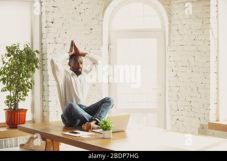 Junger afroamerikanischer Mann, der zu Hause Yoga macht, während er unter Quarantäne steht und freiberuflich online arbeitet. Remote, isoliert oder allein im Büro. Konzept des gesunden Lebensstils, Wellness, Aktivität, Bewegung. Stockfoto