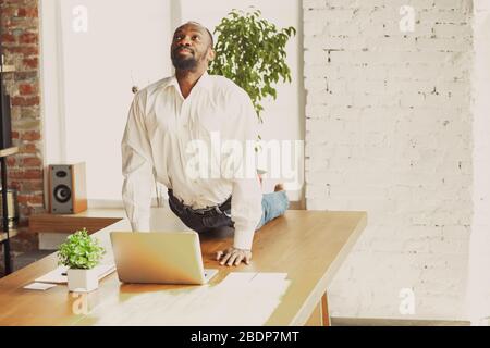 Junger afroamerikanischer Mann, der zu Hause Yoga macht, während er unter Quarantäne steht und freiberuflich online arbeitet. Remote, isoliert oder allein im Büro. Konzept des gesunden Lebensstils, Wellness, Aktivität, Bewegung. Stockfoto