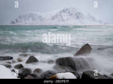 Raues Meer und Wellen, die während Schneeballzeit auf den Felsen schlagen. Entfernte schneebedeckte Berge an der Küste von Skagsanden Strand, Lofoten, Norwegen Stockfoto