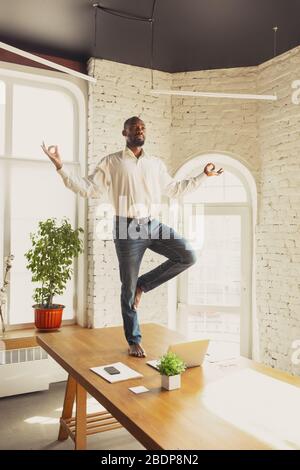 Junger afroamerikanischer Mann, der zu Hause Yoga macht, während er unter Quarantäne steht und freiberuflich online arbeitet. Remote, isoliert oder allein im Büro. Konzept des gesunden Lebensstils, Wellness, Aktivität, Bewegung. Stockfoto