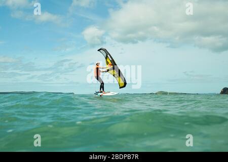 Ein Mann wingfoils in Auckland Hafen an einem Sommertag, mit einem Hand gehalten aufblasbaren Flügel und Reiten ein Tragflächenboot Surfboard. Rangitoto Island. Stockfoto