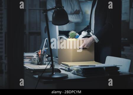 Chef feuert einen jungen Angestellten im Büro ab, sie packt ihre Sachen und hält einen Pappkarton Stockfoto