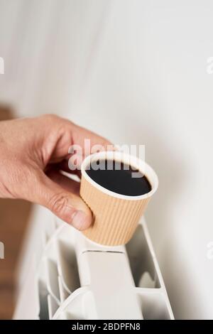 Nahaufnahme eines kaukasischen Mannes, der einen blassgrauen Pullover trägt und eine Papierbecher mit Kaffee auf einen Warmwasserkühler legt, um ihn heiß zu halten Stockfoto