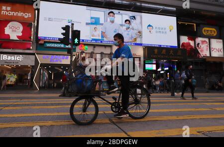 Hongkong, CHINA. April 2020. Maskierter Mann, der LP-Gas auf dem Fahrrad liefert, ist hinter ihm eine öffentliche Gesundheitswarnung, die die Bürger daran erinnert, Vorsichtsmaßnahmen gegen Infektionen mit Coronavirus zu ergreifen, die auf den großen Bildschirm projiziert werden.April-9, 2020 Hongkong.ZUMA/Liau Chung-ren Credit: Liau Chung-ren/ZUMA Wire/Alamy Live News Stockfoto