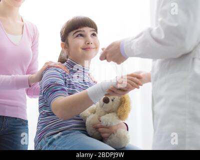 Doktor, der das verletzte Handgelenk eines Mädchens mit Gaze und Binde umhüllt, die Mutter steht neben der Tochter, erster Hilfe und Gesundheitskonzept Stockfoto