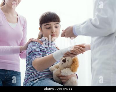 Doktor, der das verletzte Handgelenk eines Mädchens mit Gaze und Binde umhüllt, die Mutter steht neben der Tochter, erster Hilfe und Gesundheitskonzept Stockfoto