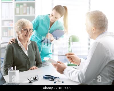 Der professionelle Arzt sitzt am Büroschreibtisch und gibt einem lächelnden älteren weiblichen Patienten ein verschreibungspflichtiges Medikament, eine Krankenschwester hält ihre Schulter, med Stockfoto