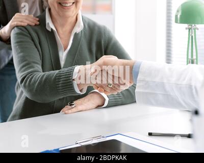 Doktor, der eine lächelnde ältere Patientin in seinem Büro begrüßt und ihre Hand schüttelt Stockfoto