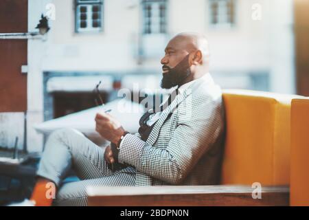 Echtes Tilt-Shift-Porträt eines schneidigen reifen kahlen afrikanischen Mannes mit einem ordentlichen Bart, in einem karierten modischen Kostüm und orangefarbenen Socken, im Freien sitzen Stockfoto