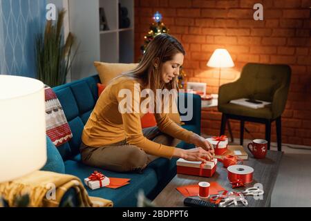 Junge Frau, die zu Hause auf dem Sofa sitzt und schöne Geschenke für Weihnachten, Feiertage und Feiern vorbereitet Stockfoto