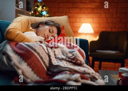 Junge Frau schläft auf dem Sofa unter einer warmen Decke, Weihnachtsbaum im Hintergrund Stockfoto