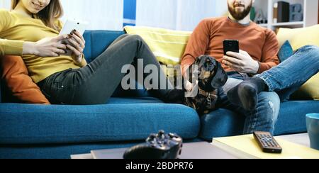Junge Paare sitzen zu Hause auf dem Sofa und verbinden sich online mit mobilen Geräten Stockfoto