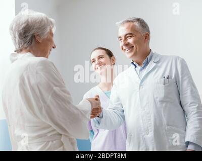 Lächelnder Facharzt und Pfleger treffen sich mit einem leitenden Patienten in der Klinik, schütteln die Hände und begrüßen sie Stockfoto