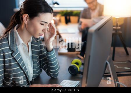 Junge, professionelle Geschäftsfrau, die am Schreibtisch sitzt und arbeitet, hat Kopfschmerzen und fühlt sich gestresst Stockfoto