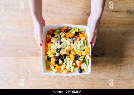 Die Hände einer Frau fangen einen Obstsalat in einer weißen Schüssel auf einem Holztisch Stockfoto