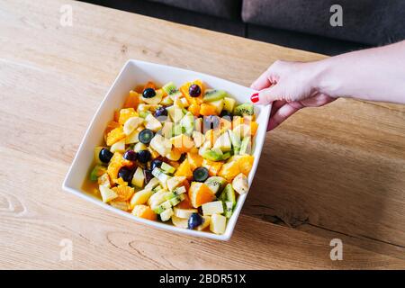 Die Hände einer Frau fangen einen Obstsalat in einer weißen Schüssel auf einem Holztisch Stockfoto