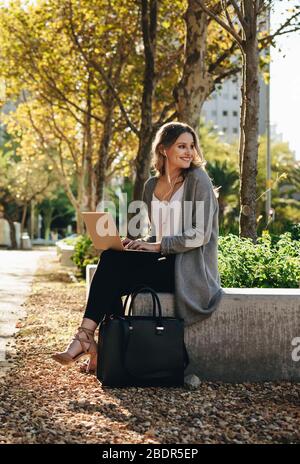 Lächelnde junge Frau, die draußen mit Laptop sitzt. Frau, die wegschaut und lächelt, während sie draußen in der Stadt sitzt. Stockfoto