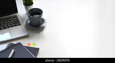 Foto von Computer-Laptop, Kaffeetasse, Notebook, Tagebuch, Stift, Topfpflanze setzen auf weißen Schreibtisch. Geordnetes Arbeitsplatzkonzept. Stockfoto